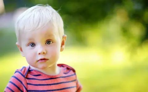 Ein kleiner Junge mit blonden Haaren und blauen Augen blickt neugierig in die Kamera. Sein gestreiftes Shirt erinnert an die Vielfalt von Jungennamen international.