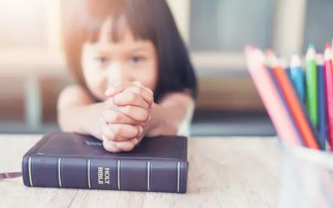 Ein kleines Mädchen betet mit gefalteten Händen über einer Bibel. Vielleicht lernt sie gerade etwas über biblische Frauennamen. Buntstifte liegen daneben.