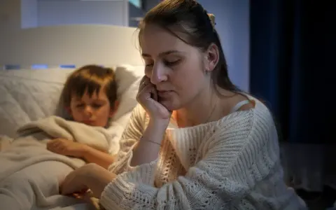Müde Mutter sitzt neben ihrem schlafenden Sohn, der immer beim Aufwachen weint. Sie hält ihren Kopf in der Hand.