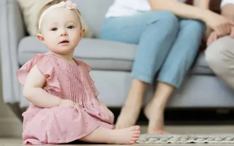 Ein süßes Baby in rosa Kleidchen, mit einem Blumenband im Haar, sitzt auf dem Boden. Im Hintergrund sitzen Eltern. Vielleicht heißt sie Zofia oder Hanna, typisch polnische vornamen weiblich.