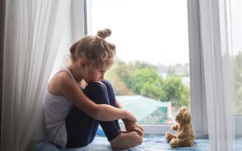 Ein kleines Mädchen sitzt traurig am Fenster, ihr Teddybär neben ihr. Sie denken über den Tod mit Kindern besprechen nach.