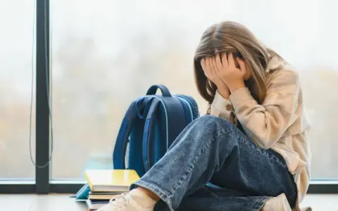Ein Mädchen mit verdecktem Gesicht sitzt neben einem Rucksack und Büchern. Sie leidet unter **Angst vor Schule ohne Grund**.
