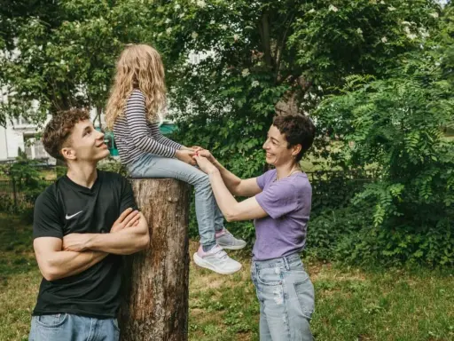 Ein junger Mann schaut zu seiner Mutter, die mit seiner kleinen Schwester auf einem Baumstumpf sitzt. Vielleicht denkt er, wenn der Sohn die falsche Freundin hat, ist die Familie trotzdem da.