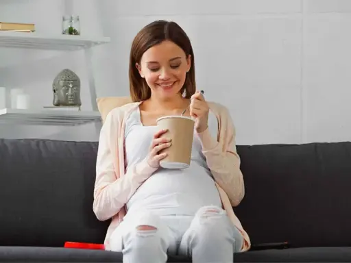 Lächelnde Schwangere isst Eis auf dem Sofa. Ein süßer Genuss während der Eis Schwangerschaft.