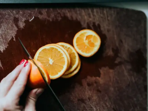 Eine Hand mit roten Nägeln schneidet Orangen auf einem Holzbrett. Orangen in der Schwangerschaft sind eine gesunde Wahl.