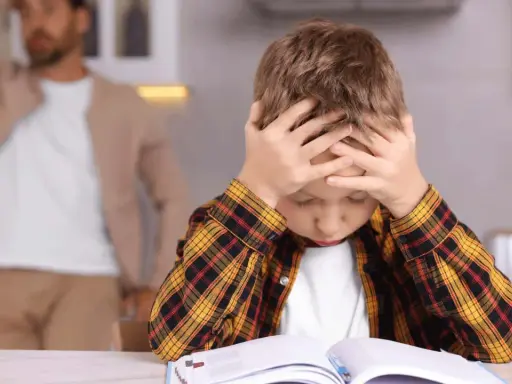 Junge mit Händen am Kopf, der frustriert seine Hausaufgaben macht. Vater im Hintergrund.