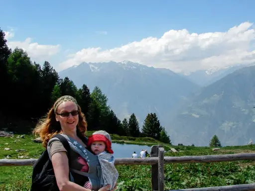 Frau mit Baby im Tragetuch genießt die Berglandschaft. Ein Beispiel für alleine reisen mit Kind.