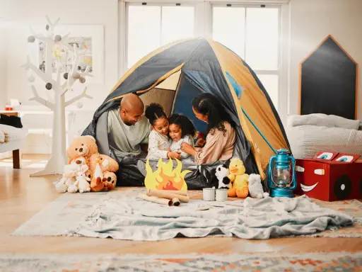 Familie verbringt **gemeinsame Zeit** beim Zelten im Wohnzimmer. Kinder kuscheln mit Teddybären, während Eltern lachen.