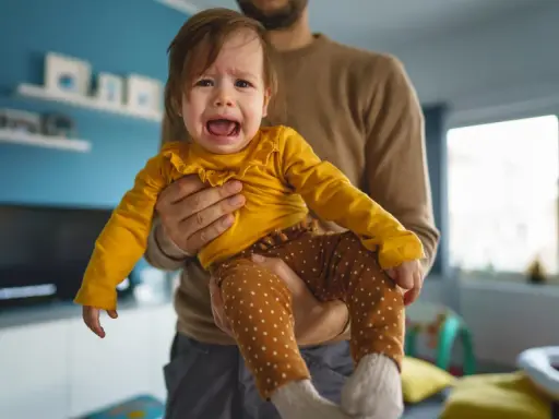 Ein weinendes Baby in gelbem Oberteil und gepunkteter Hose wird von seinem Vater gehalten. Typische "Mama-Phase".