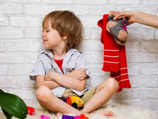 Ein Junge sitzt mit verschränkten Armen und einem trotzigen Gesichtsausdruck da. Über ihm hält eine Hand einen roten Schal und ein Paar Socken. Der Junge will sich nicht anziehen.