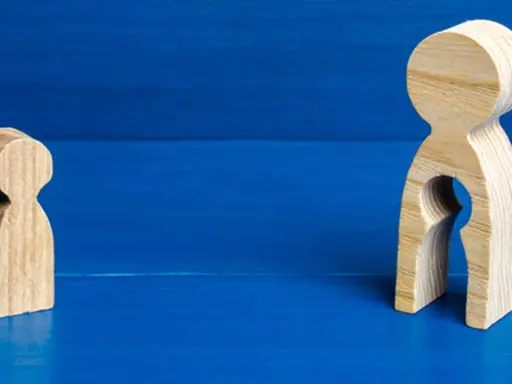 Zwei Holzfiguren auf blauem Grund. Eine kleine Figur steht allein. Die größere Figur hat eine leere Öffnung, die an ein Kind erinnert, und symbolisiert das Bedauern über Mutterschaft.