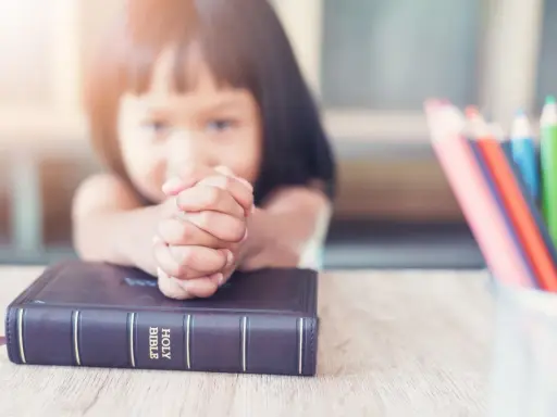 Ein kleines Mädchen betet mit gefalteten Händen über einer Bibel. Vielleicht lernt sie gerade etwas über biblische Frauennamen. Buntstifte liegen daneben.