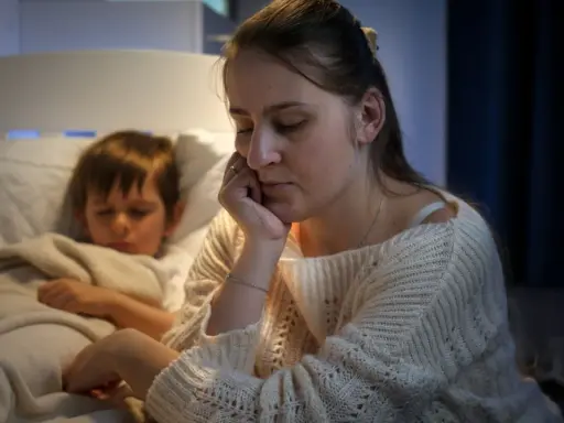 Müde Mutter sitzt neben ihrem schlafenden Sohn, der immer beim Aufwachen weint. Sie hält ihren Kopf in der Hand.