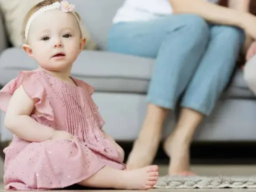 Ein süßes Baby in rosa Kleidchen, mit einem Blumenband im Haar, sitzt auf dem Boden. Im Hintergrund sitzen Eltern. Vielleicht heißt sie Zofia oder Hanna, typisch polnische vornamen weiblich.