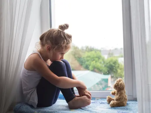 Ein kleines Mädchen sitzt traurig am Fenster, ihr Teddybär neben ihr. Sie denken über den Tod mit Kindern besprechen nach.