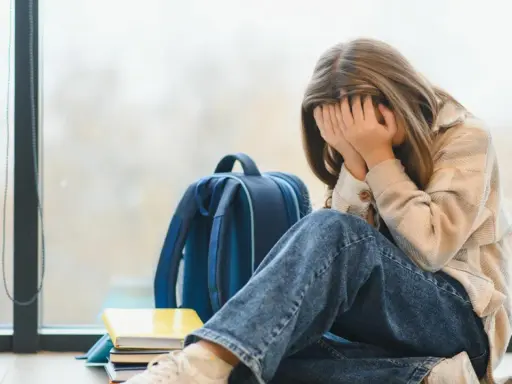 Ein Mädchen mit verdecktem Gesicht sitzt neben einem Rucksack und Büchern. Sie leidet unter **Angst vor Schule ohne Grund**.