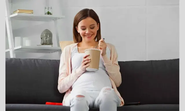 Lächelnde Schwangere isst Eis auf dem Sofa. Ein süßer Genuss während der Eis Schwangerschaft.