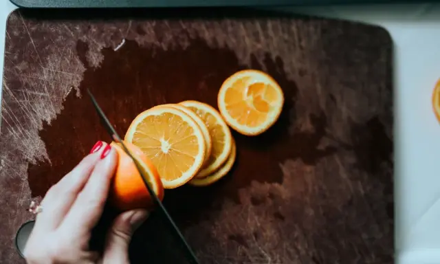 Eine Hand mit roten Nägeln schneidet Orangen auf einem Holzbrett. Orangen in der Schwangerschaft sind eine gesunde Wahl.