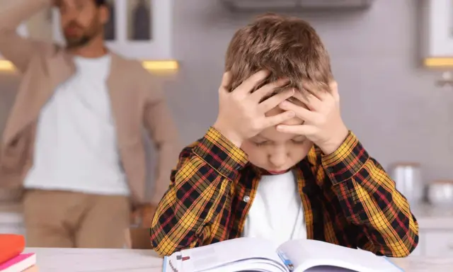 Junge mit Händen am Kopf, der frustriert seine Hausaufgaben macht. Vater im Hintergrund.