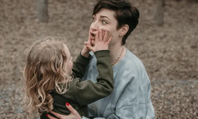 Ein kleines Kind drückt der Mutter spielerisch die Hände ins Gesicht. Vielleicht fragt es sich, warum beleidigen Kinder andere Kinder, wenn sie doch so viel Spaß haben können.