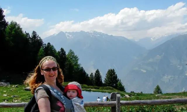 Frau mit Baby im Tragetuch genießt die Berglandschaft. Ein Beispiel für alleine reisen mit Kind.