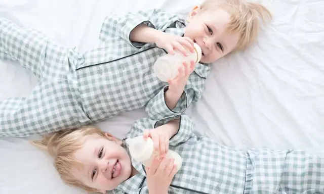 Zwei süße Jungen in karierten Pyjamas trinken aus Fläschchen. Perfekt für Inspiration für Zwillingsnamen Jungen!