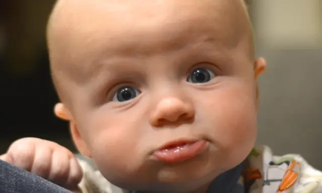 Nahaufnahme eines Babys mit großen blauen Augen und einem lustigen Gesichtsausdruck. Das Baby scheint sich selbst zu erkunden, seine Hand ruht auf einer dunklen Oberfläche.