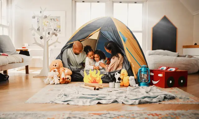 Familie verbringt **gemeinsame Zeit** beim Zelten im Wohnzimmer. Kinder kuscheln mit Teddybären, während Eltern lachen.