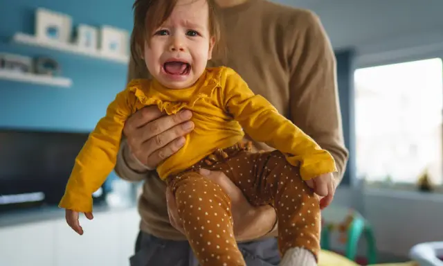 Ein weinendes Baby in gelbem Oberteil und gepunkteter Hose wird von seinem Vater gehalten. Typische "Mama-Phase".