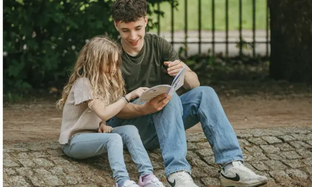 Ein junger Mann liest einem Mädchen im Park vor. Trotz der Trennung der Eltern finden die Kinder Trost und Nähe in gemeinsamen Momenten.