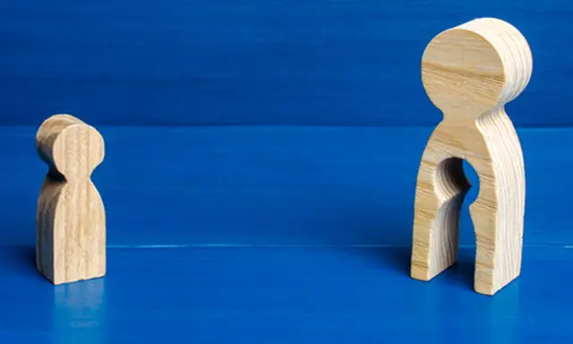 Zwei Holzfiguren auf blauem Grund. Eine kleine Figur steht allein. Die größere Figur hat eine leere Öffnung, die an ein Kind erinnert, und symbolisiert das Bedauern über Mutterschaft.