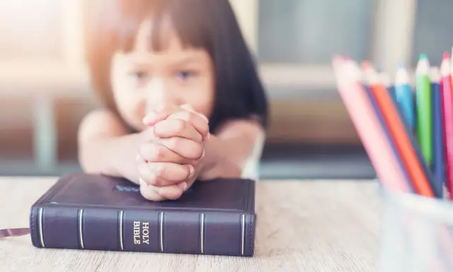 Ein kleines Mädchen betet mit gefalteten Händen über einer Bibel. Vielleicht lernt sie gerade etwas über biblische Frauennamen. Buntstifte liegen daneben.