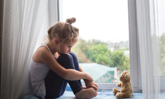 Ein kleines Mädchen sitzt traurig am Fenster, ihr Teddybär neben ihr. Sie denken über den Tod mit Kindern besprechen nach.