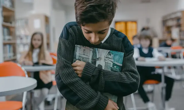 Ein Junge hält ein Buch, während andere Kinder im Hintergrund spielen. Er wirkt traurig, als ob mein Kind im Kindergarten ausgeschlossen wird.