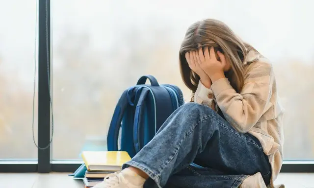 Ein Mädchen mit verdecktem Gesicht sitzt neben einem Rucksack und Büchern. Sie leidet unter **Angst vor Schule ohne Grund**.