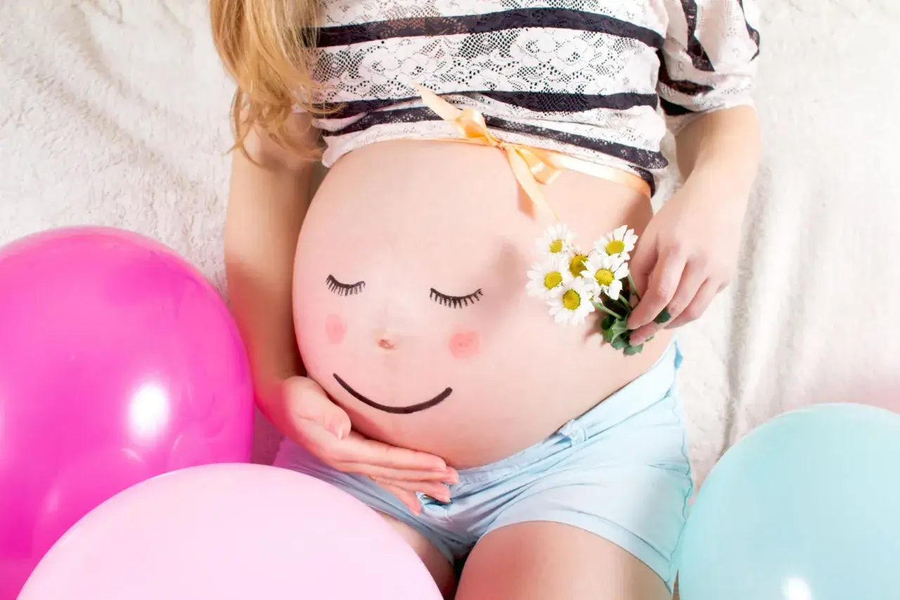 Ein bemalter Babybauch mit einem lächelnden Gesicht, umgeben von bunten Ballons und Gänseblümchen. Ein süßer Moment des Babybauch bemalen.