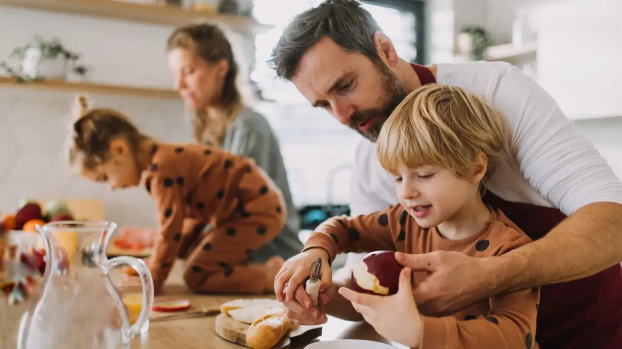 Vater hilft Sohn beim Schälen eines Apfels. Die Familie bereitet gemeinsam das Essen vor, ein Beispiel für geteilten mental load.