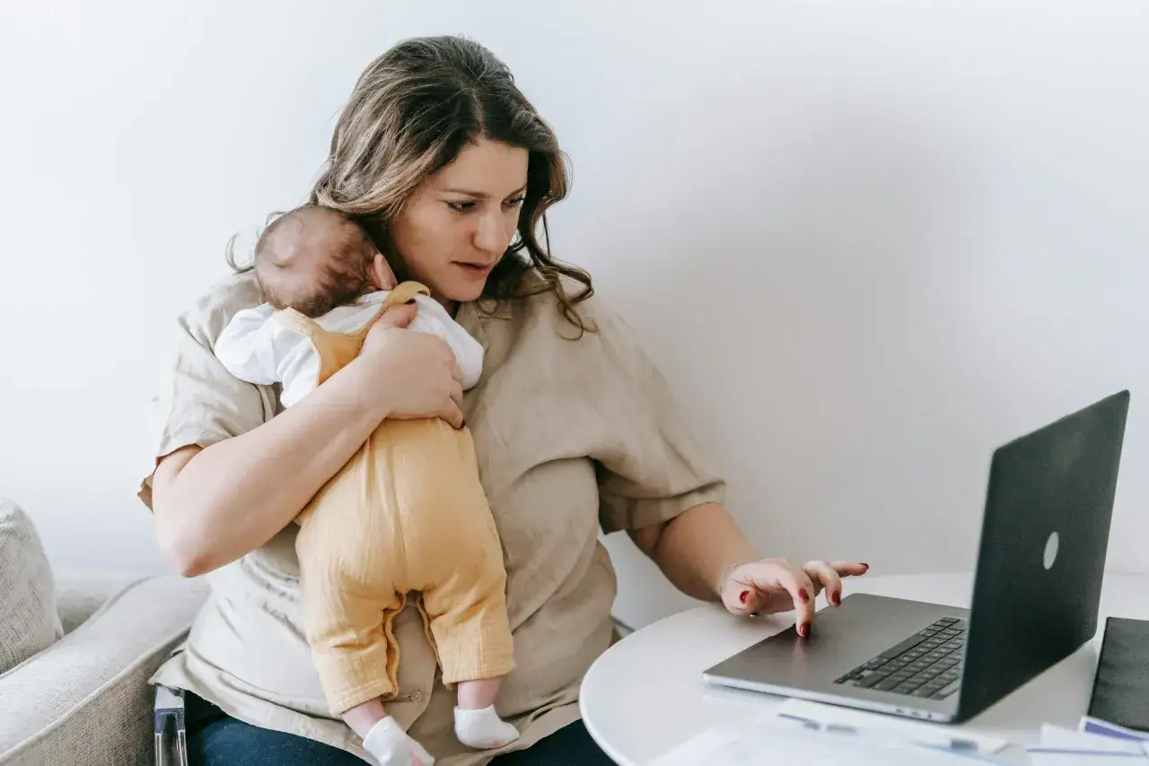 Mutter mit Baby auf dem Arm blickt nachdenklich auf Laptop. M&ouml;gliche Symptome von 