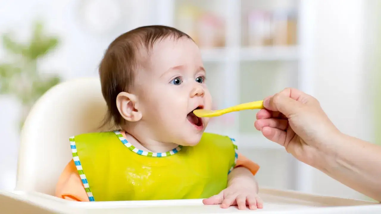 Baby mit Lätzchen bekommt Brei auf Löffel. Reifezeichen Baby: offener Mund, neugieriger Blick.