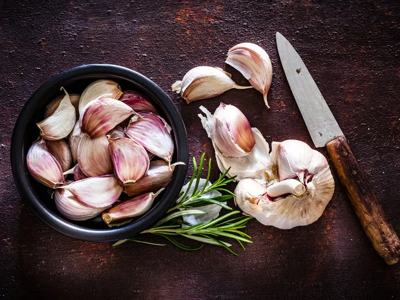Knoblauch und Rosmarin auf dunklem Untergrund. Ein Messer liegt daneben. Knoblauch in der Schwangerschaft kann ein n&uuml;tzliches Gew&uuml;rz sein.
