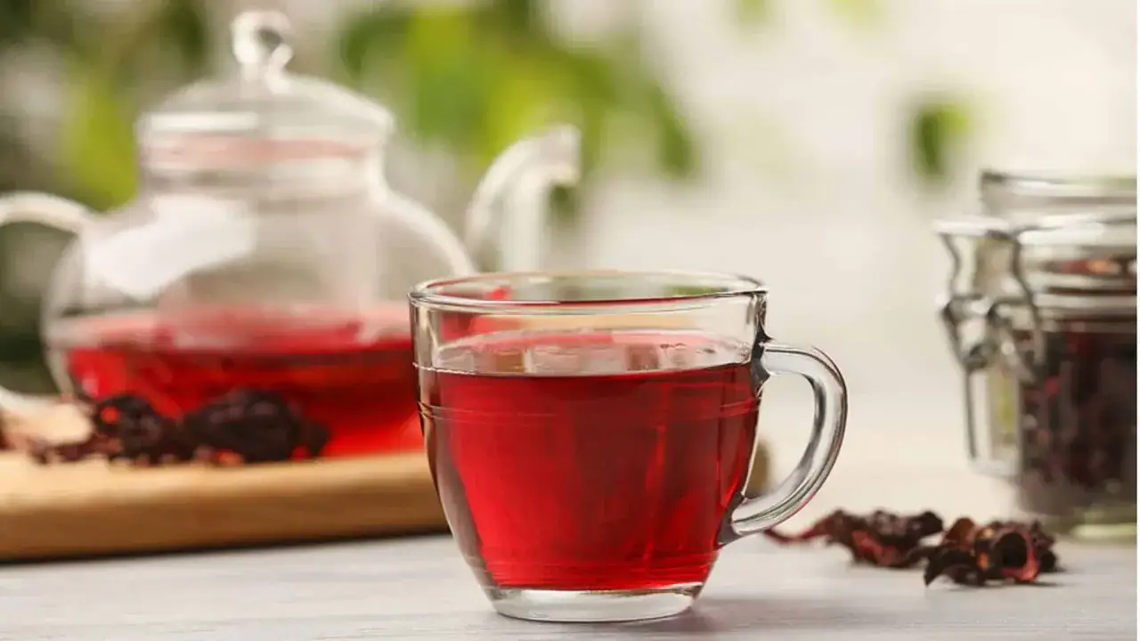 Ein Glas leuchtend roter Hibiskustee, perfekt für die Stillzeit. Daneben eine Teekanne und getrocknete Hibiskusblüten.