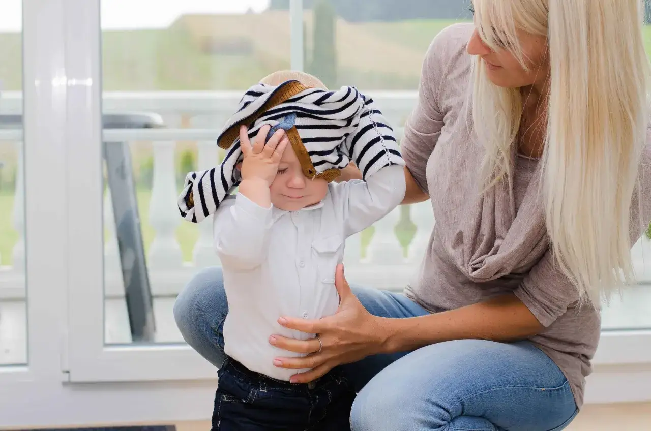 Mutter versucht, ihrem Kind ein gestreiftes Oberteil &uuml;ber den Kopf zu ziehen, aber das Kind will sich nicht anziehen und h&auml;lt sich die Stirn.