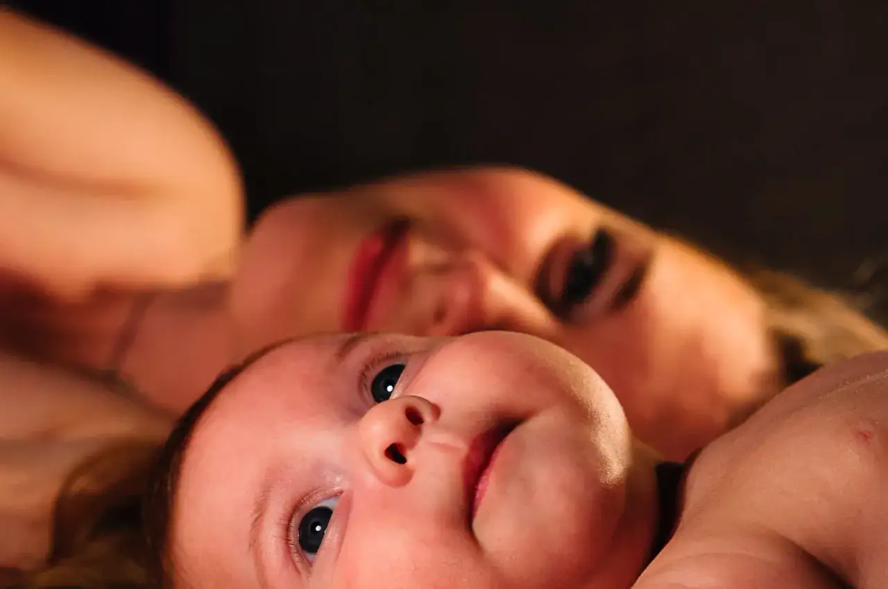 Ein Baby im REM-Schlaf, mit weit ge&ouml;ffneten Augen, blickt neugierig in die Kamera, w&auml;hrend im Hintergrund die unscharfe Silhouette einer Frau zu sehen ist.