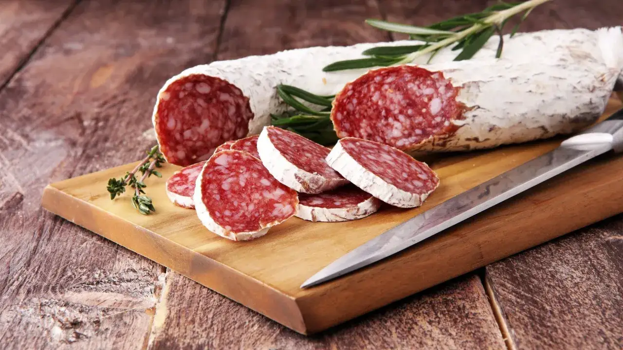 Salami auf einem Holzbrett, daneben ein Messer und Kr&auml;uter. Darf man in der Schwangerschaft Salami essen? Diese Frage besch&auml;ftigt viele werdende M&uuml;tter.
