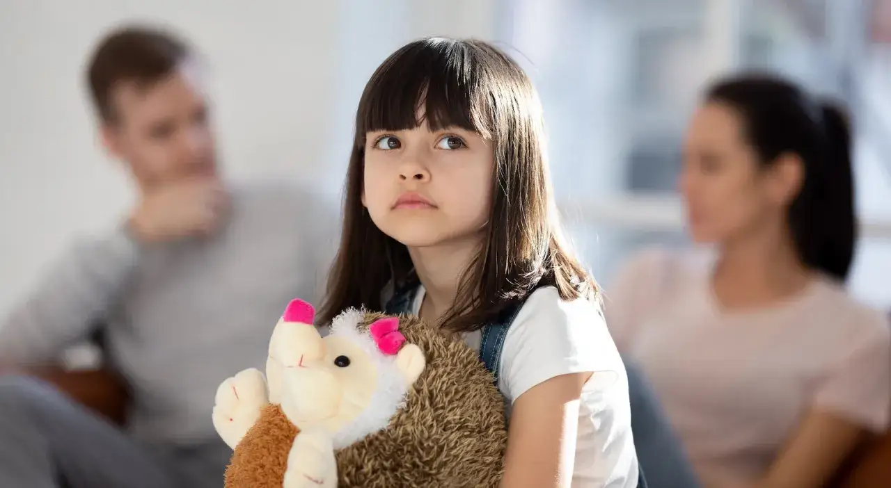 Ein trauriges Mädchen hält seinen Teddybär fest, während im Hintergrund die Eltern, die sich trennen, zu sehen sind.