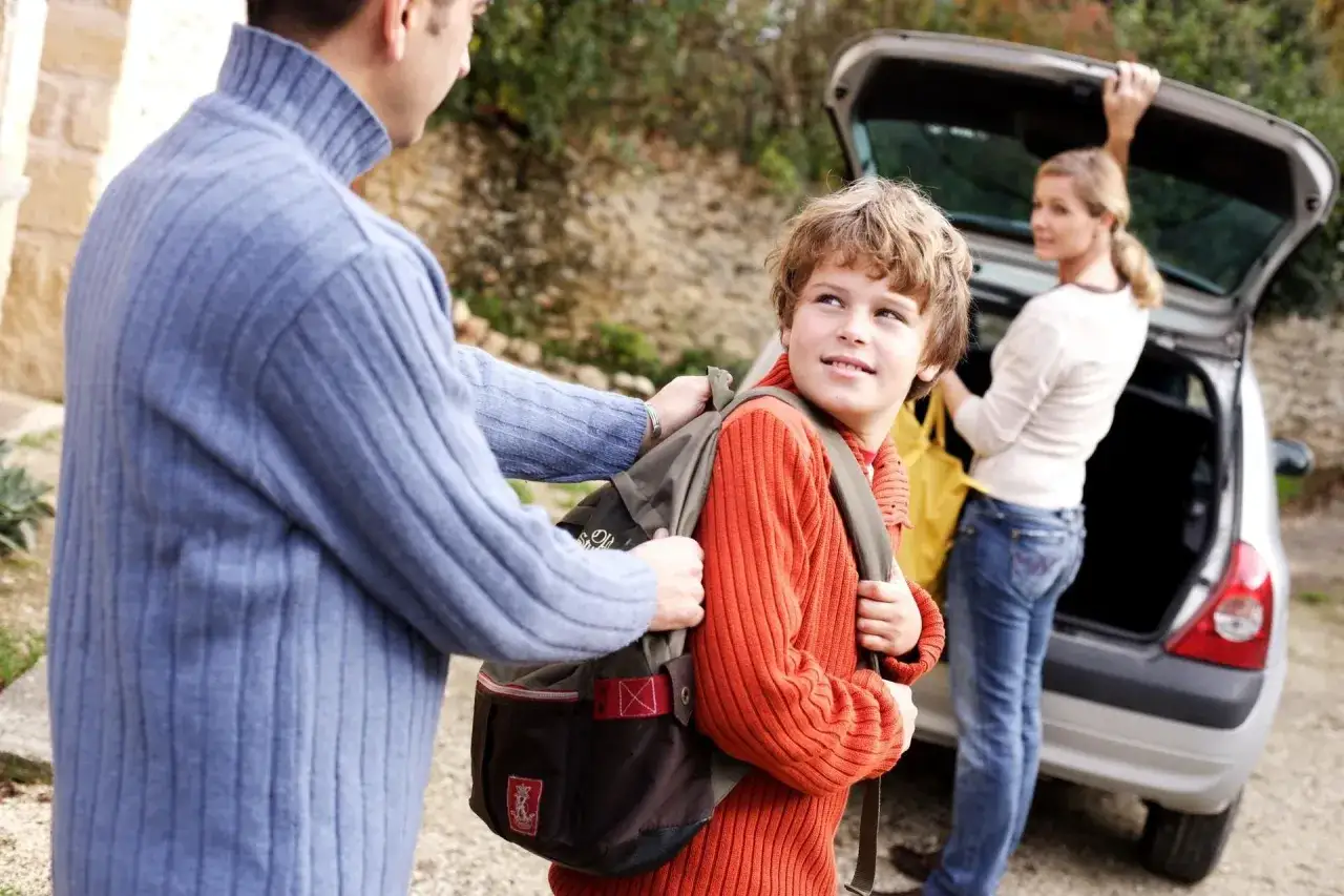 Vater hilft Sohn mit Rucksack, Mutter bel&auml;dt Auto. Ein Beispiel f&uuml;r die praktische Umsetzung der gesetzlichen Regelung Kinderbetreuung Trennung.