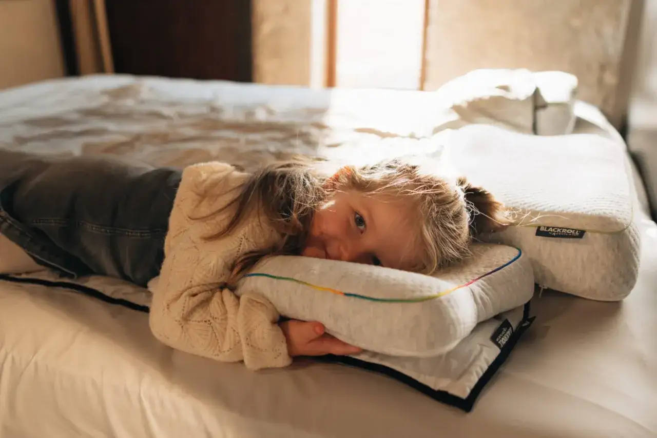 Ein kleines Mädchen kuschelt sich auf einem Bett mit einem bunten Kissen. Sie scheint bereit, alleine ins Bett zu gehen, und genießt die Gemütlichkeit.