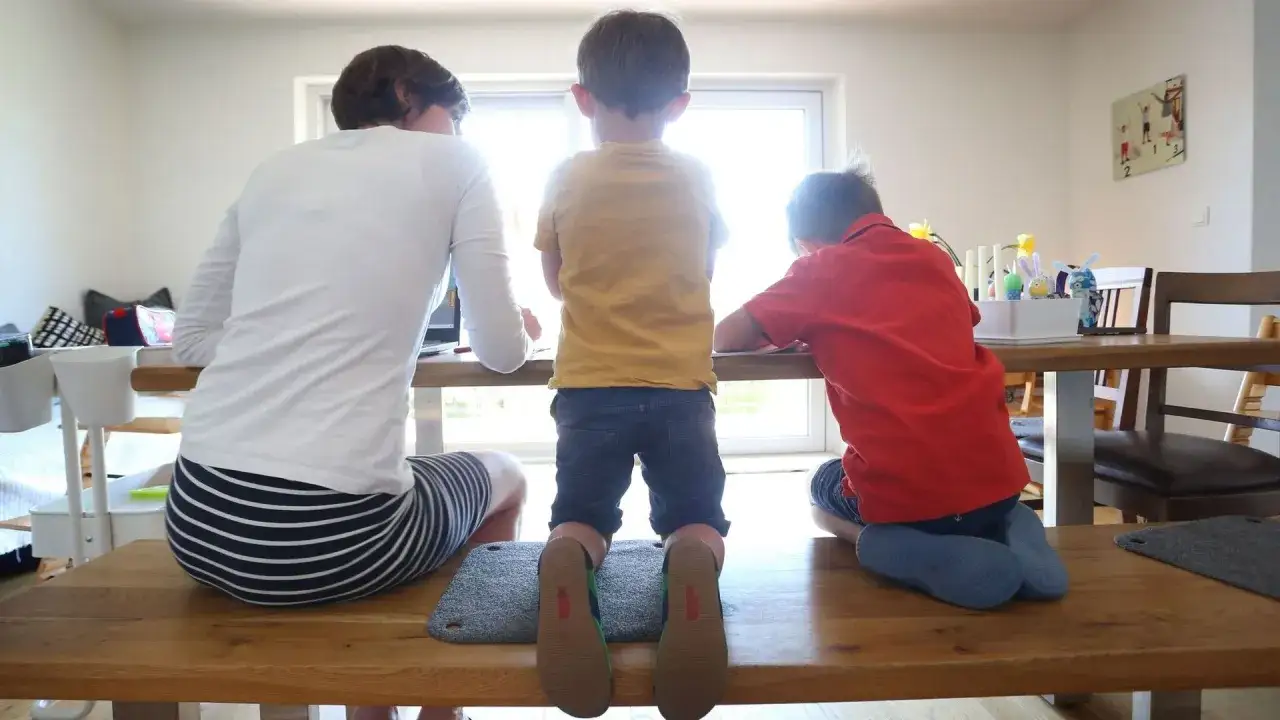 Eine Frau und zwei Kinder sitzen an einem Tisch. Die Frau ist mit ihrer Arbeit besch&auml;ftigt, w&auml;hrend die Kinder malen. Die Szene k&ouml;nnte eine toxische Tochter darstellen, die ihre Mutter ignoriert.