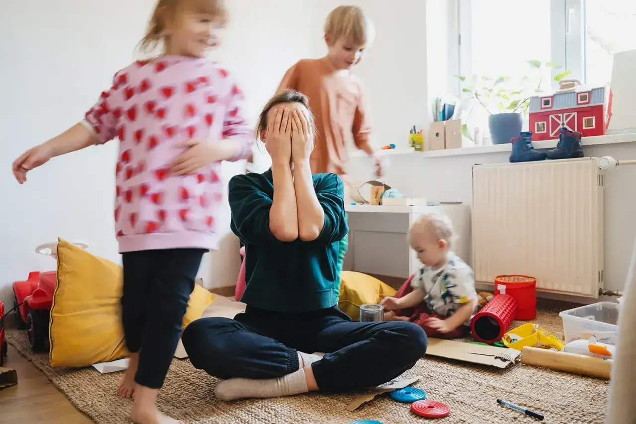 Mutter schimpft mit Kind. Sie h&auml;lt sich die H&auml;nde vor das Gesicht, w&auml;hrend die Kinder im Hintergrund spielen.