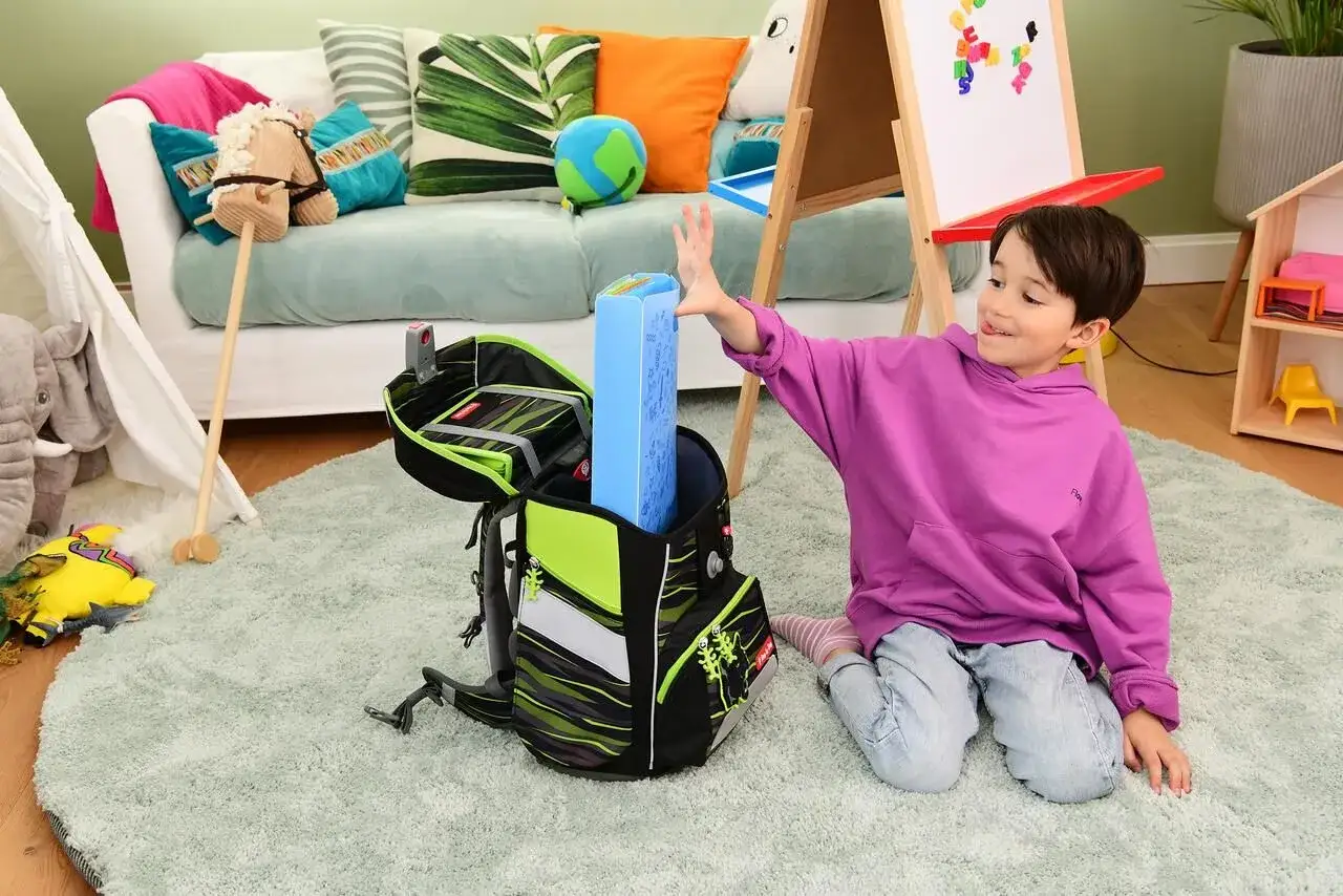 Junge packt Schulranzen f&uuml;r einen guten Schulstart nach den Ferien. Bunter Ranzen, Spielzeug und Tafel im Hintergrund.